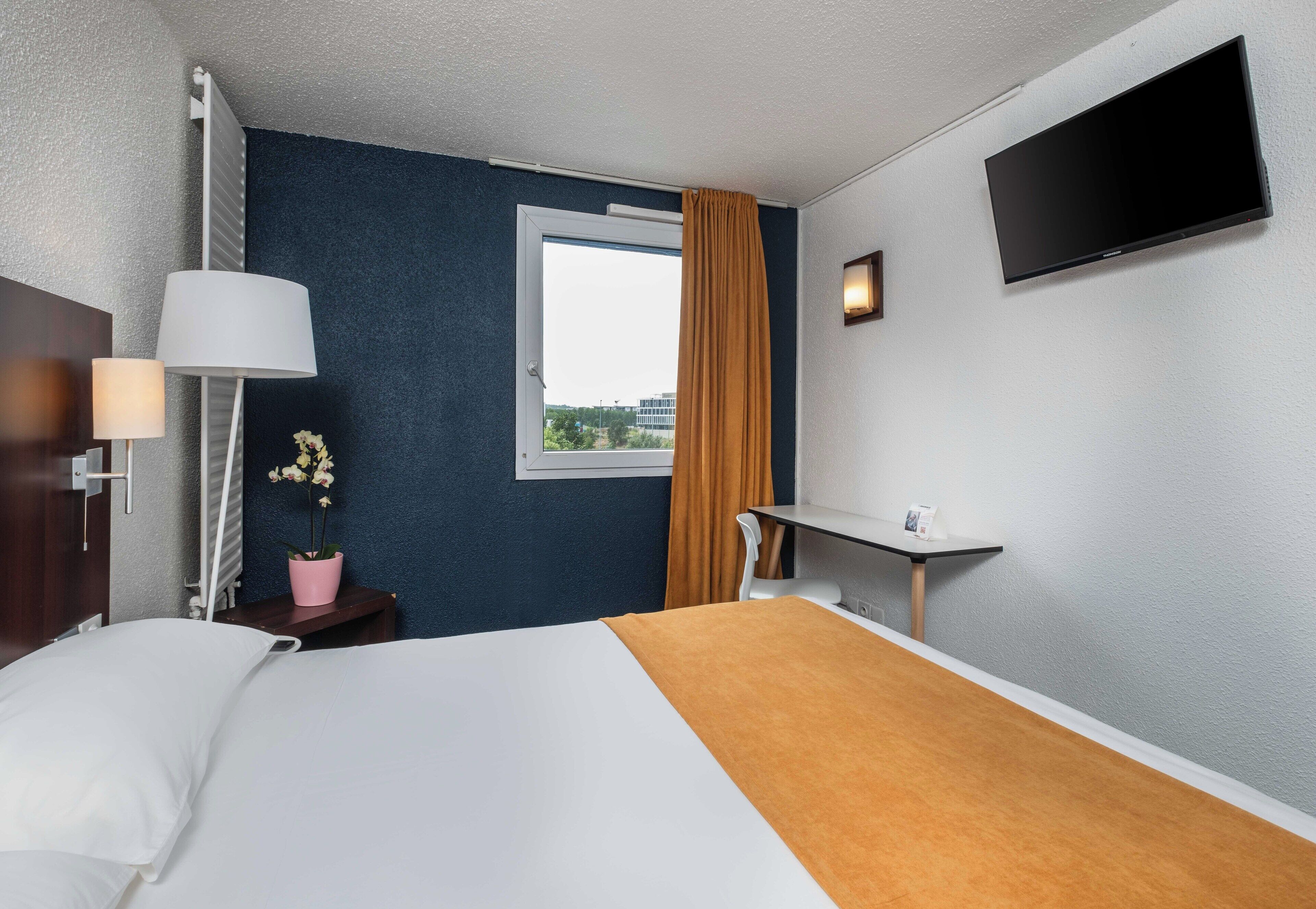Ett hotellrum med en säng, ett skrivbord, en tv, ett fönster med gardiner och en krukväxt.