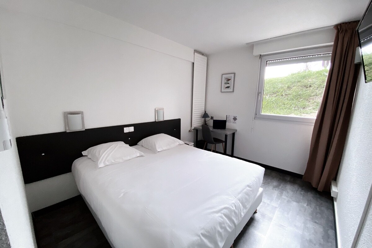 Une chambre d’hôtel avec un lit, un bureau avec un ordinateur portable, une fenêtre avec des rideaux et un lampadaire fixé au mur.