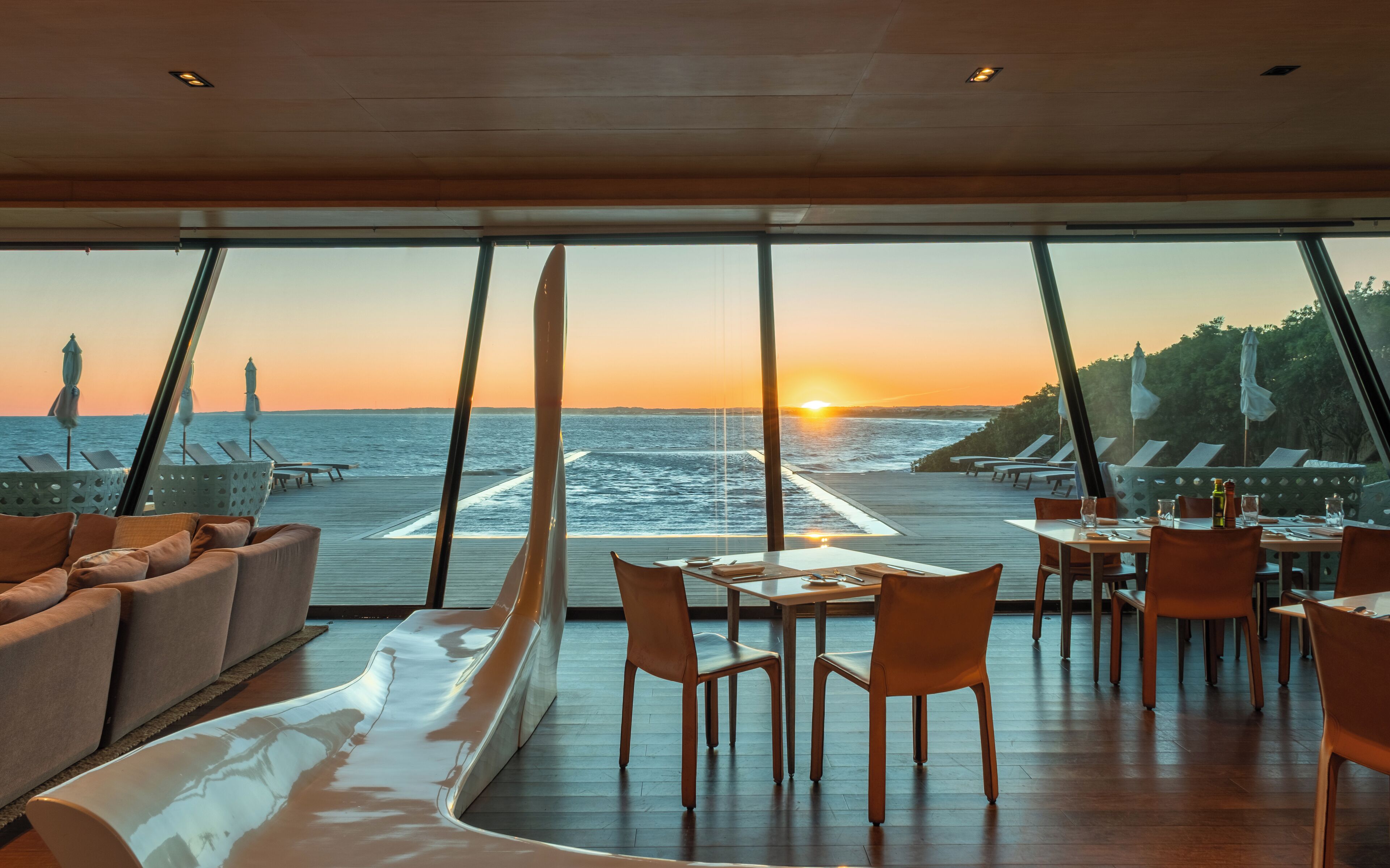 Un espace repas avec des tables et des chaises, un canapé et une vue sur l’océan au coucher du soleil.
