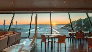 Un comedor con mesas y sillas, un sofá y vista al océano al atardecer.