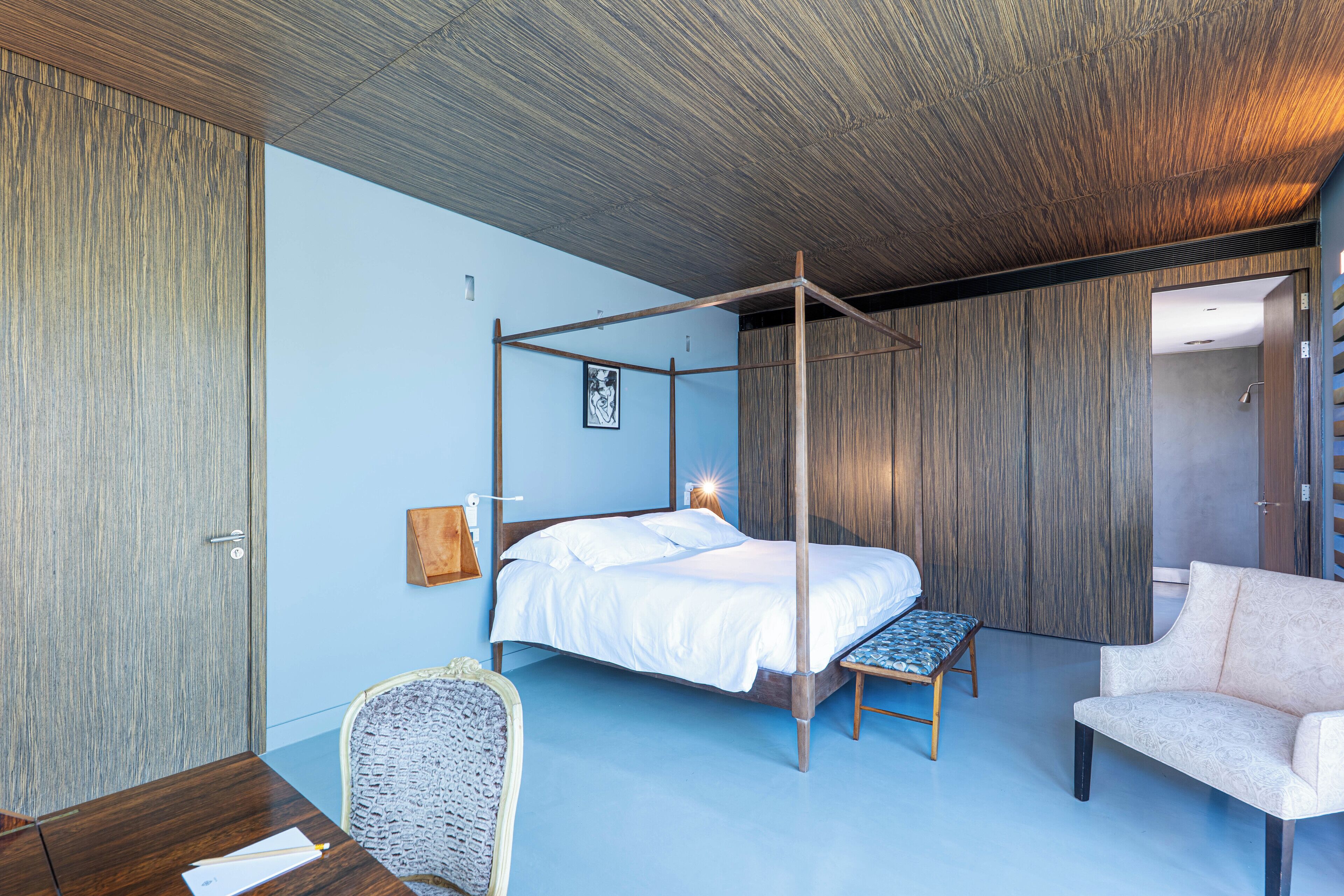 A modern bedroom with a four-poster bed, a wooden ceiling, and a blue wall.