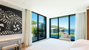 A modern hotel room with a large bed, a desk, and a view of a coastal landscape through floor-to-ceiling windows. - Playa Vik Jose Ignacio (José Ignacio)