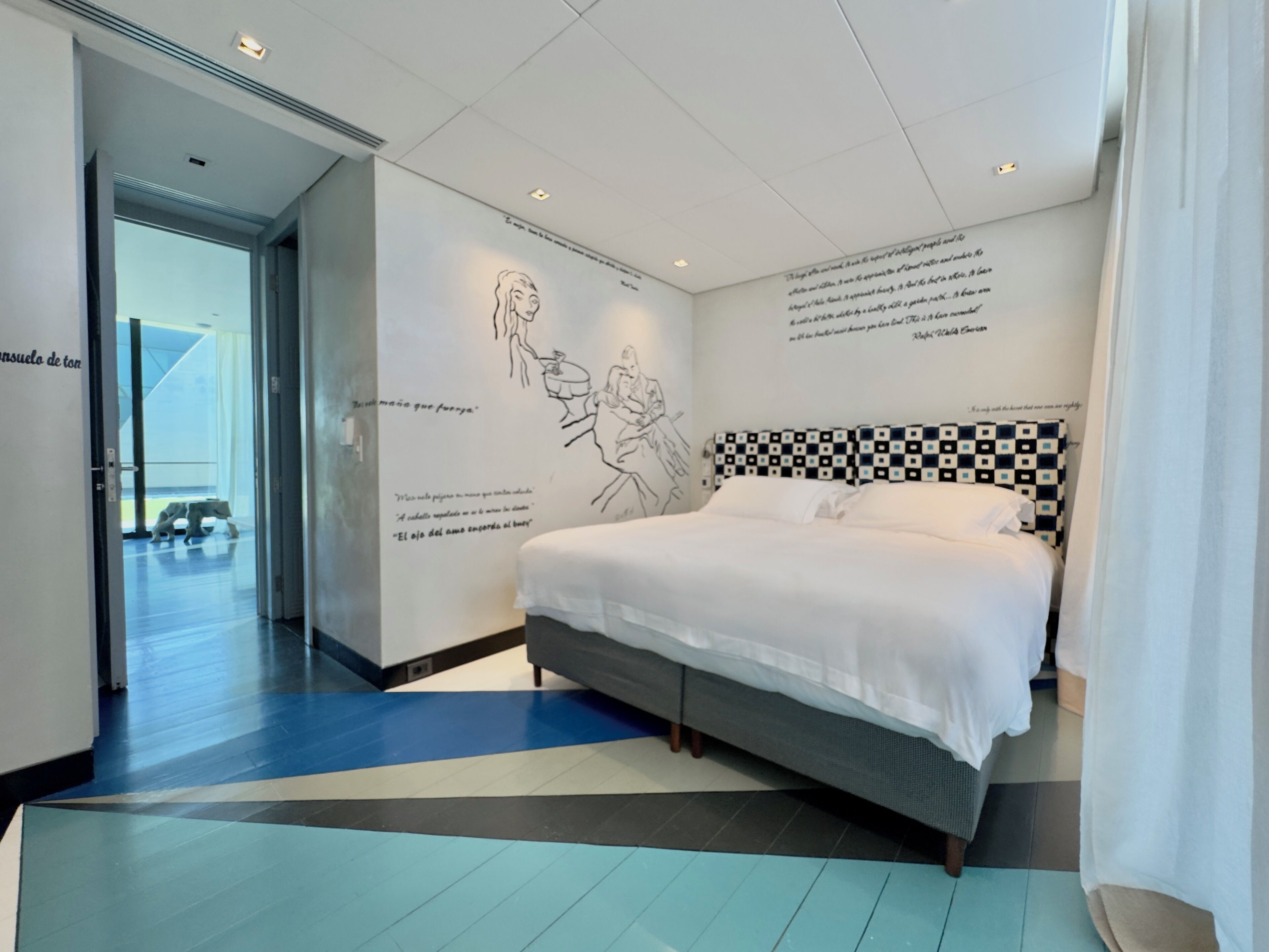 Quarto de hotel moderno com uma cama grande, cabeceira xadrez e paredes decoradas com esboços e citações em preto e branco.