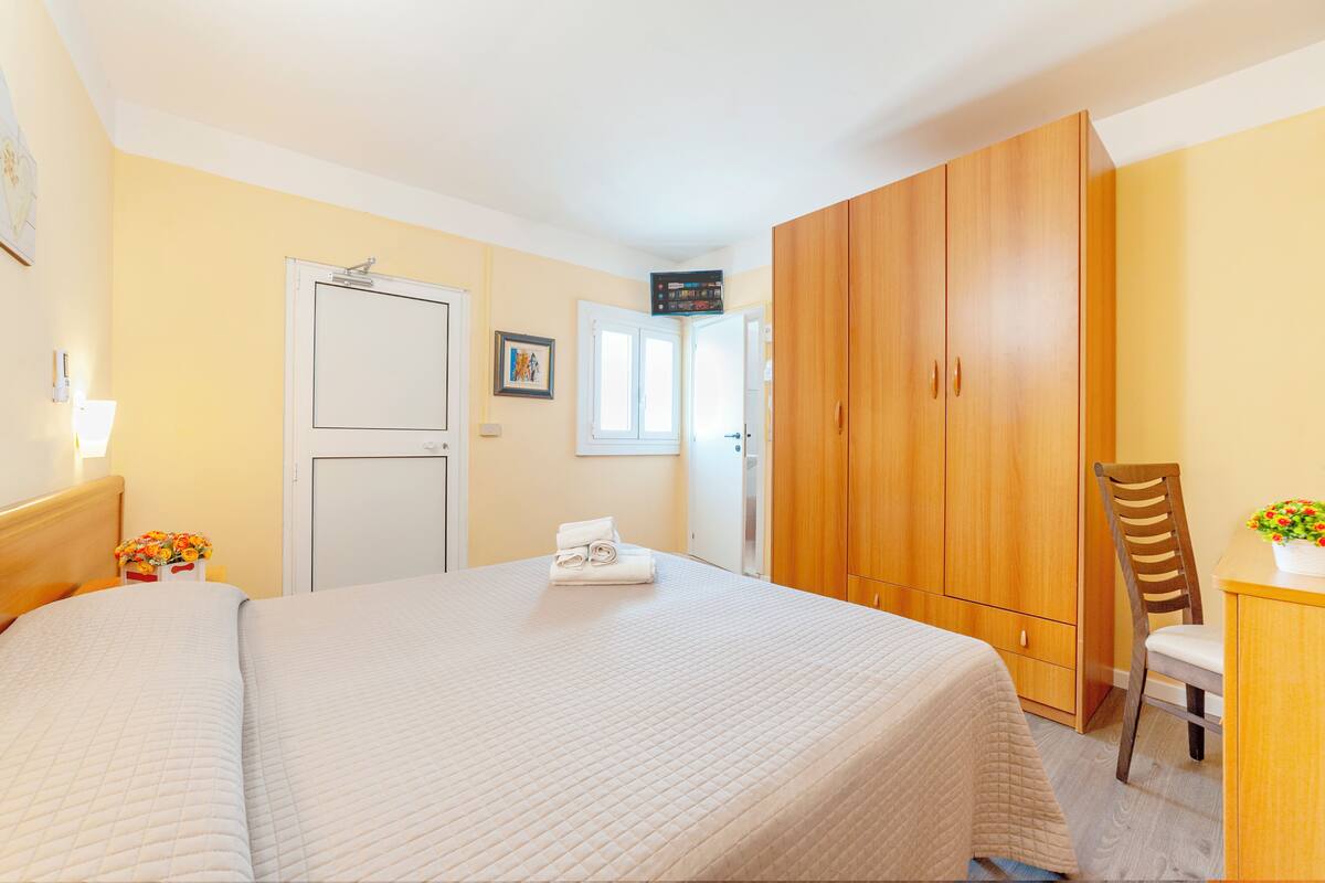 Ein Hotelzimmer mit einem großen Bett, einem Kleiderschrank, einem Schreibtisch mit Stuhl und einem kleinen Tisch mit Blumen.