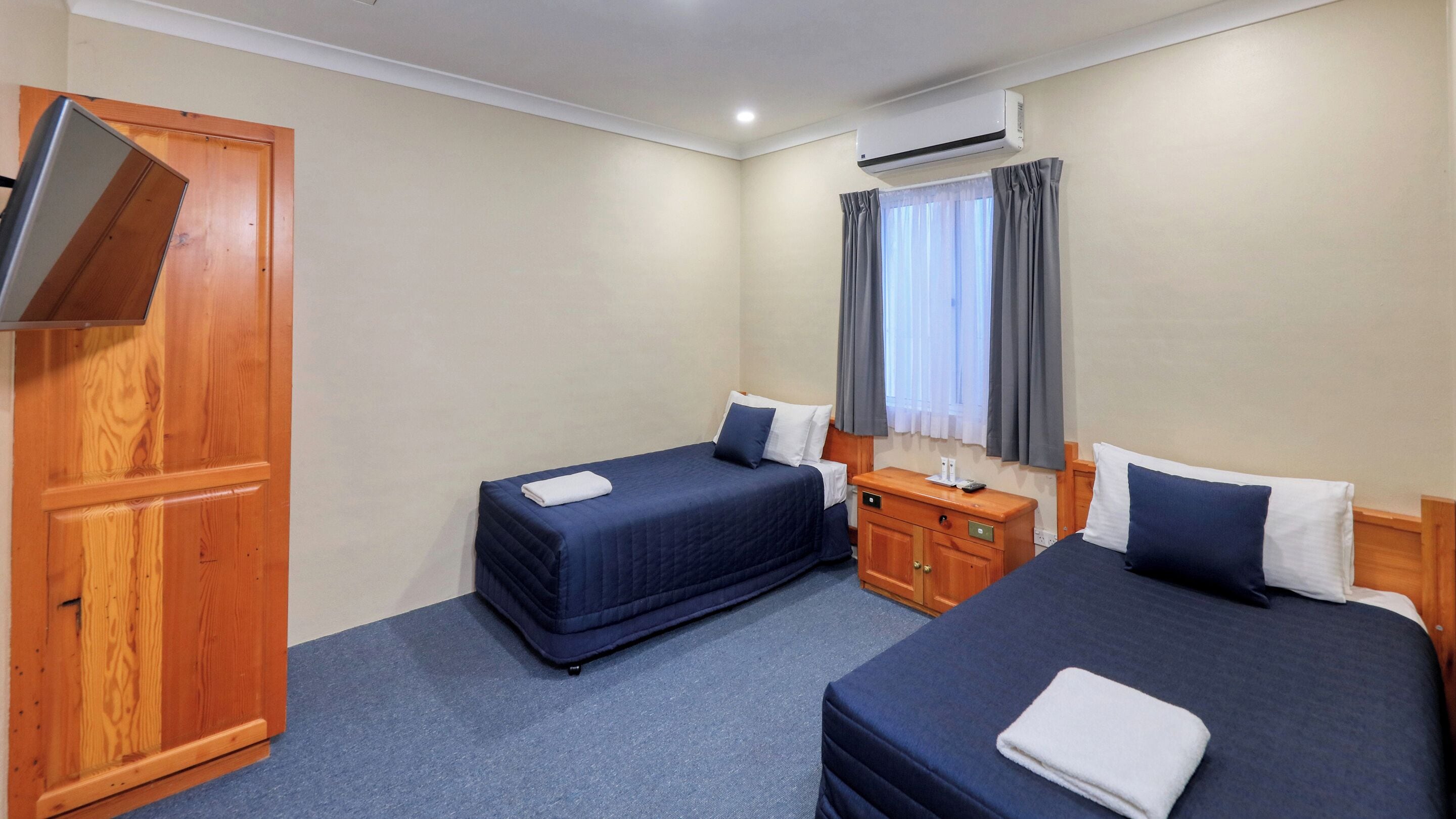A hotel room with two beds, a wardrobe, a bedside table, and an air conditioning unit.