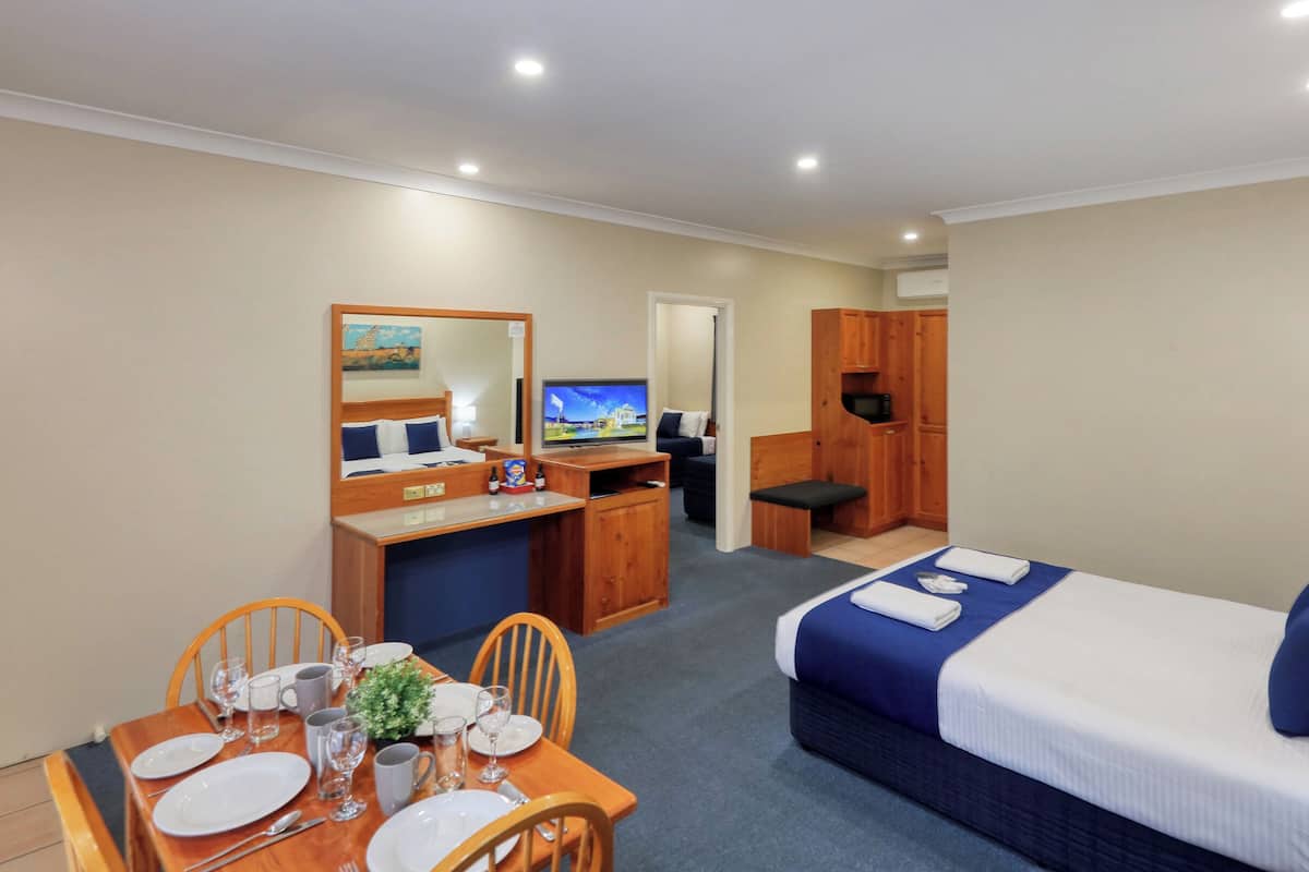 A hotel room with a bed, a desk with a computer, a TV, a dining area with a table set for two, and a small kitchenette.