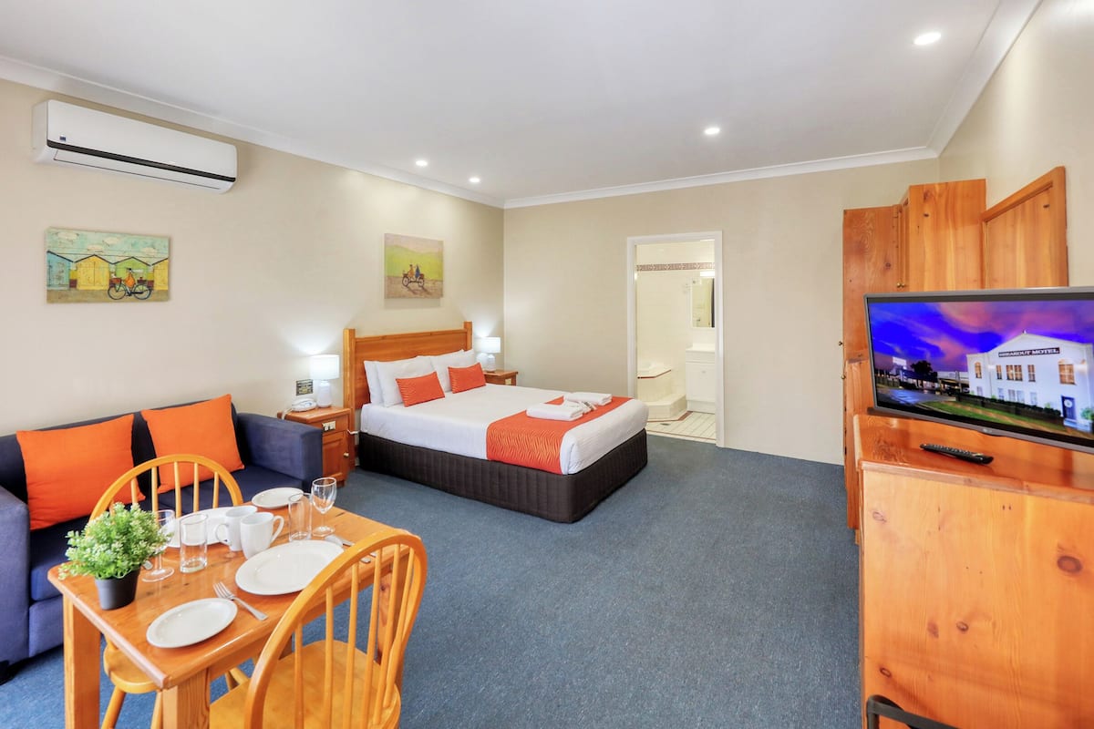 A hotel room with a bed, a sofa, a dining table with chairs, a TV, and an air conditioning unit.