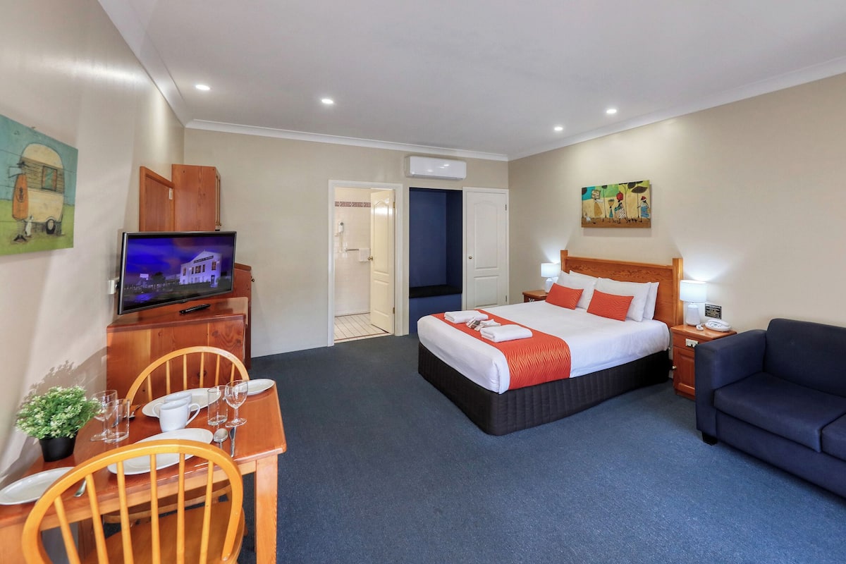 A hotel room with a bed, a TV, a desk with a chair, a sofa, and a painting on the wall.