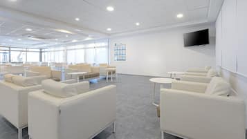 A modern, well-lit lounge area with white sofas and tables, large windows, and a mounted TV on the wall.