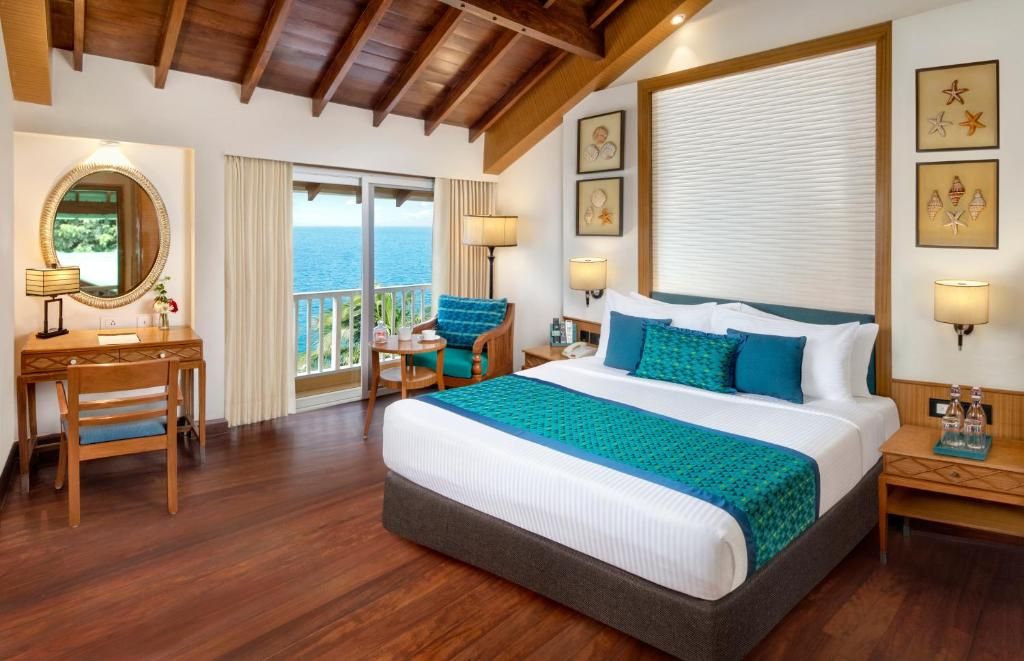 A hotel room with a large bed, a desk, a chair, a balcony with a view of the sea, and artwork on the walls.