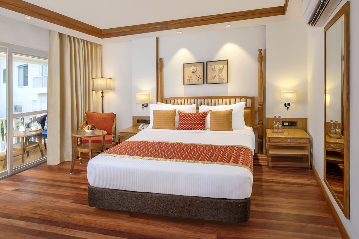 Ein Hotelzimmer mit einem großen Bett, einem Holz-Kopfende, zwei Wandleuchten, einem kleinen Tisch mit einer Lampe, einem Sessel und einem Balkon mit Blick auf Gebäude und Grünflächen.