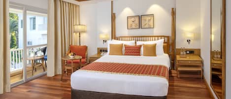 Habitación de hotel con una cama grande, cabecera de madera, dos lámparas fijadas a la pared, una mesita con lámpara, una silla y un balcón con vistas a edificios y vegetación.