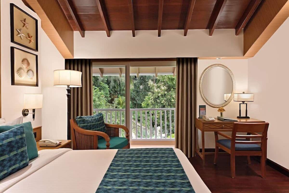 Ein Hotelzimmer mit Bett, Schreibtisch, Stuhl, Spiegel und einem Balkon mit Blick auf Grünflächen.