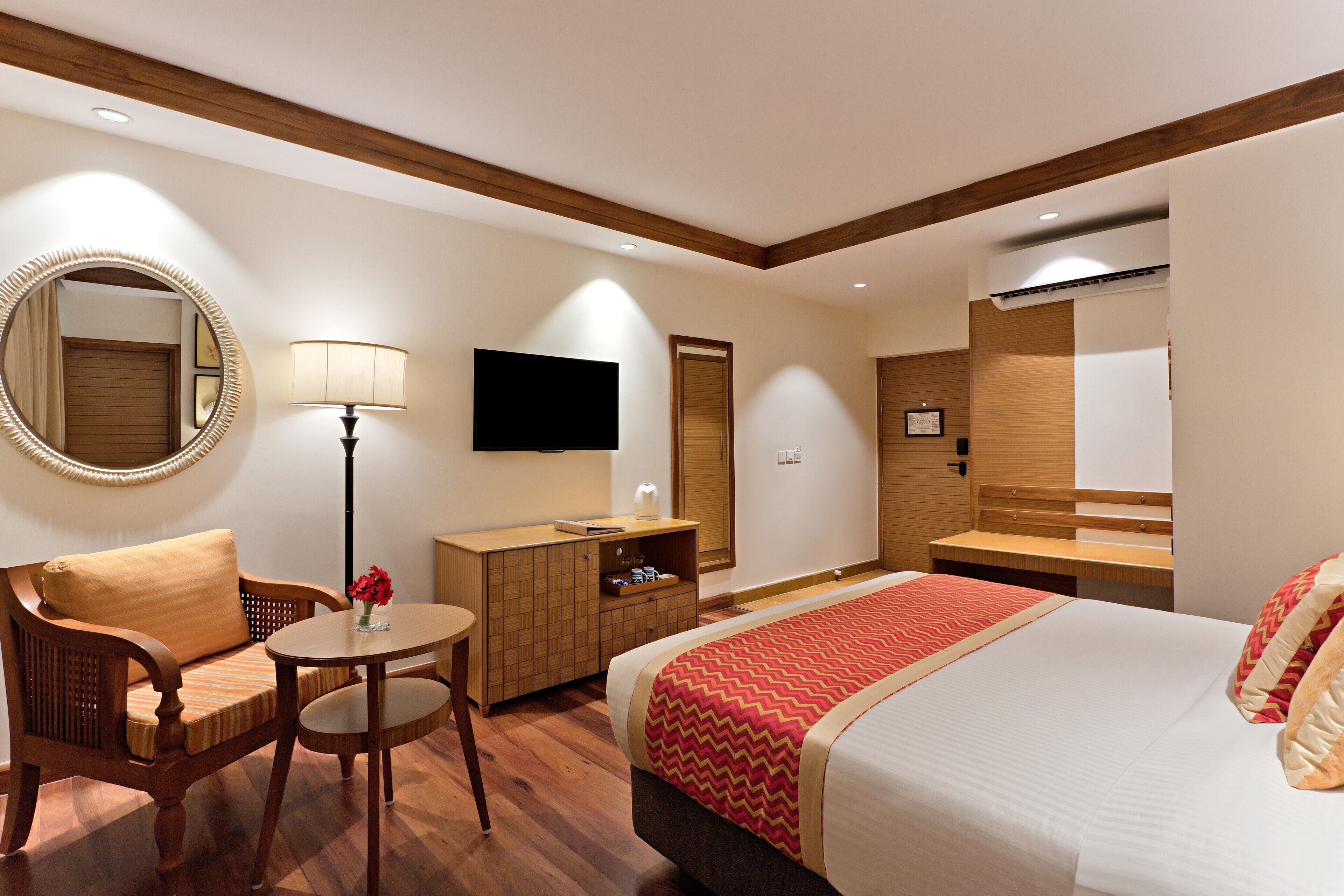 A hotel room with a bed, a wooden chair, a small table with a lamp, a TV, and a mirror.