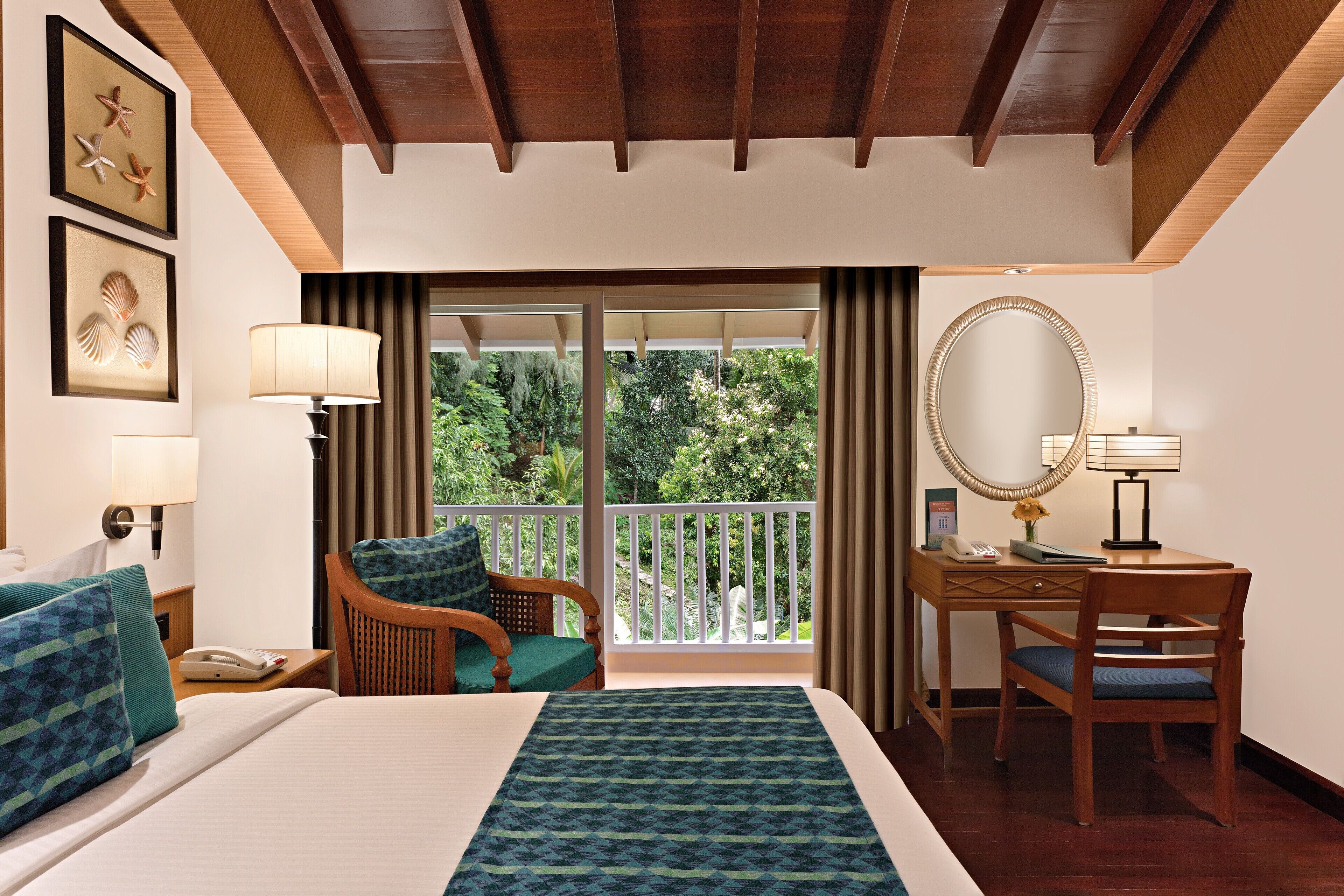 A hotel room with a bed, a desk with a chair, a large mirror, and a balcony with a view of greenery.