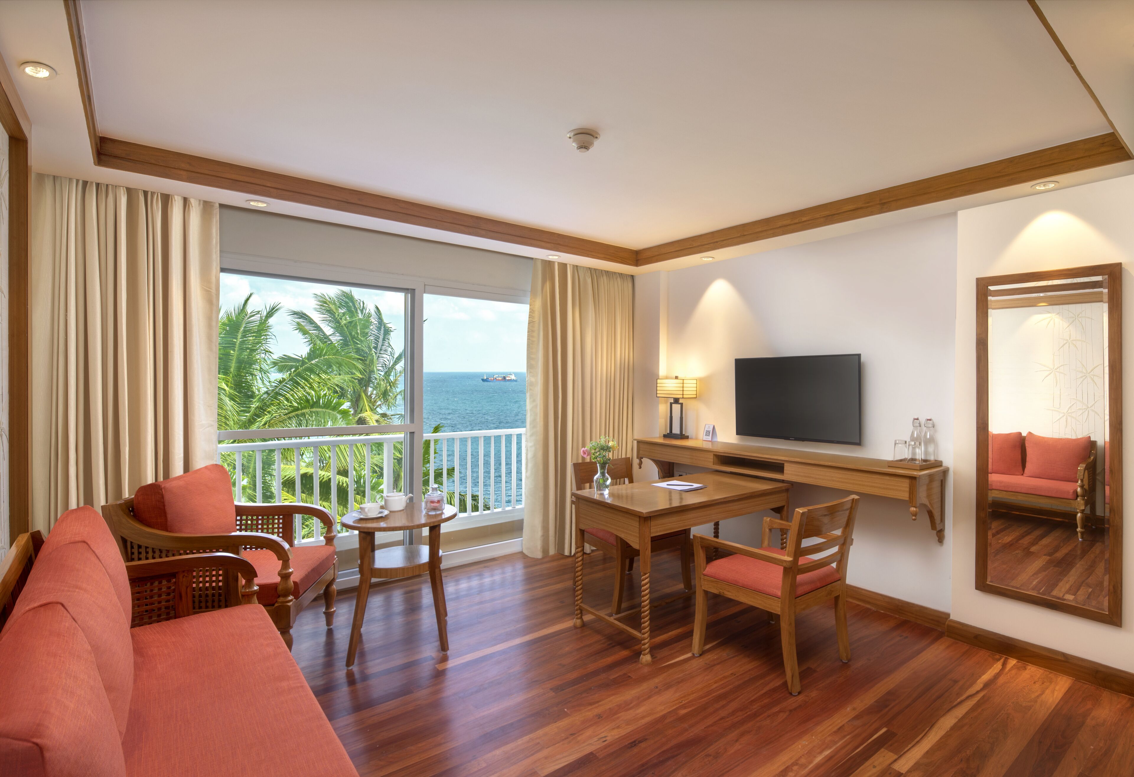 Une chambre d’hôtel avec un plancher en bois, un canapé, une table à manger avec des chaises, un téléviseur posé sur un meuble, un balcon avec vue sur l’océan et un grand miroir au mur.