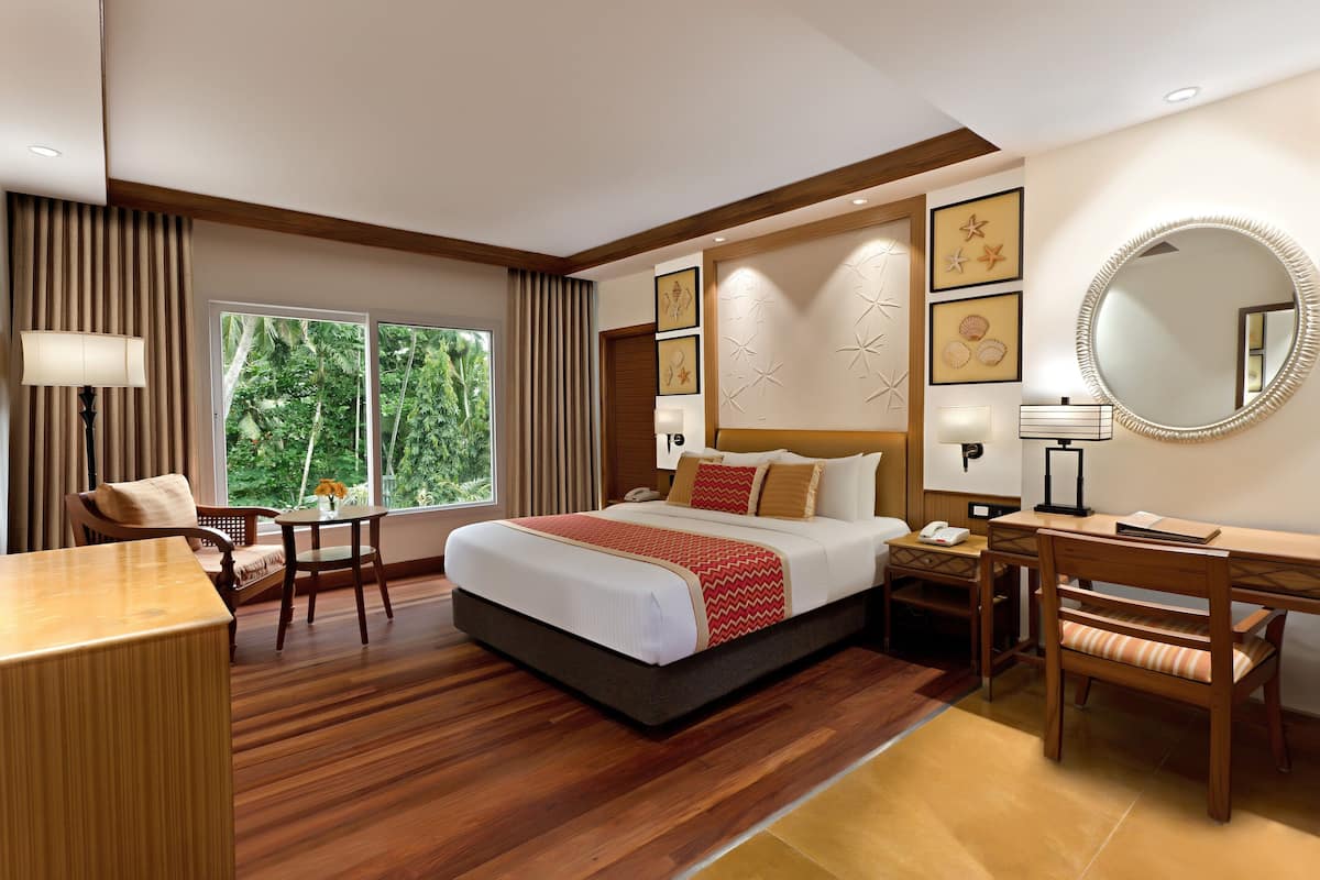 Ein Hotelzimmer mit einem großen Bett, einem Schreibtisch, einem Stuhl, einem Schminktisch mit Spiegel und einem Fenster mit Blick auf Grünflächen.