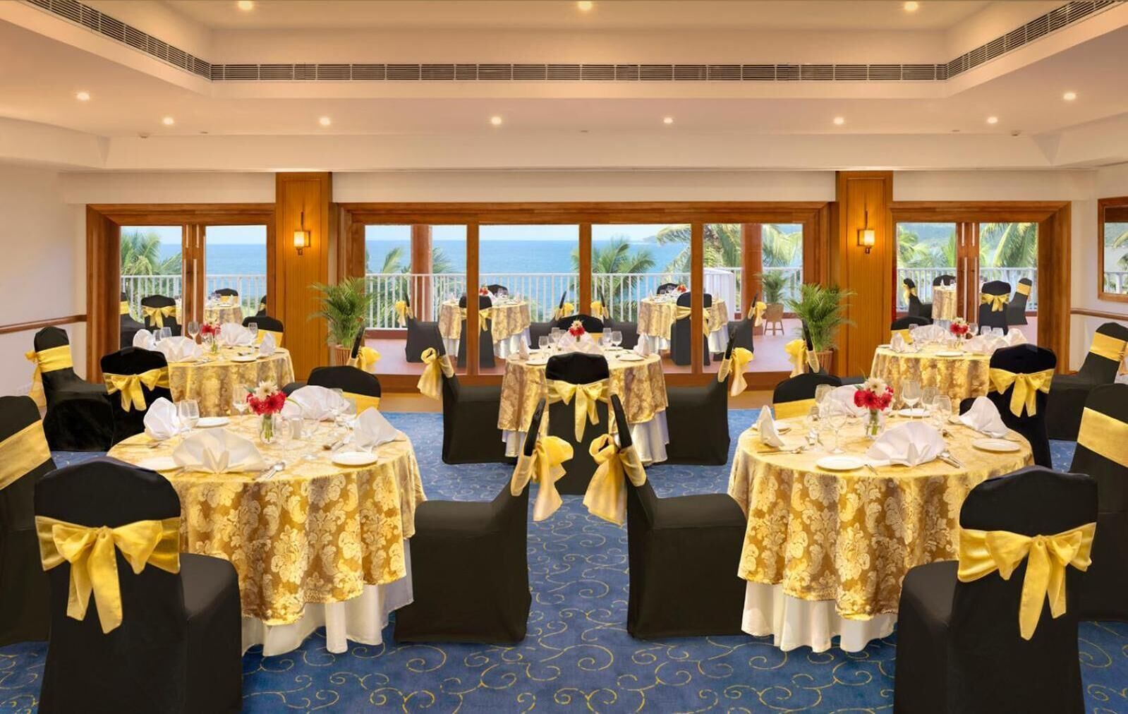 A banquet hall with round tables, black chairs with yellow sashes, and a view of the ocean through large windows.