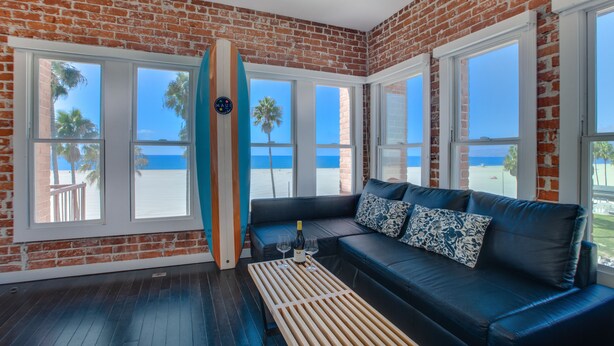 Una habitación con pared de ladrillo, ventanas amplias, sofá de cuero azul y una tabla de surf.
