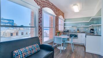 A modern kitchen with a dining area, a black leather sofa with a patterned pillow, and large windows offering a view of the city.