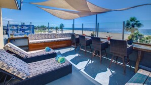 Un área de estar al aire libre iluminada por el sol, con vistas a la playa, que cuenta con muebles de mimbre y un banco de madera.