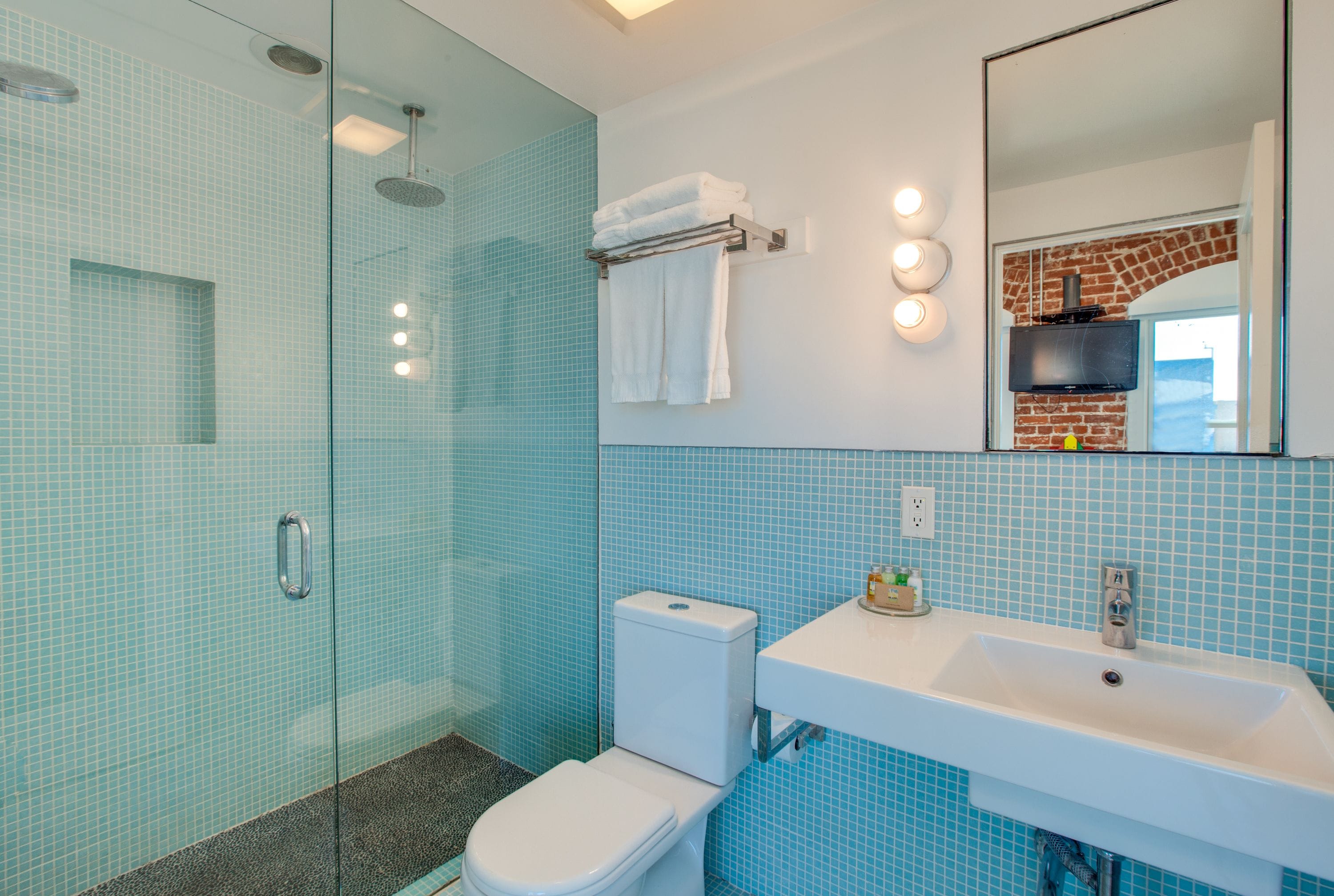 A bathroom with a glass shower enclosure, a white toilet, a sink with a mirror above it, and blue-tiled walls.