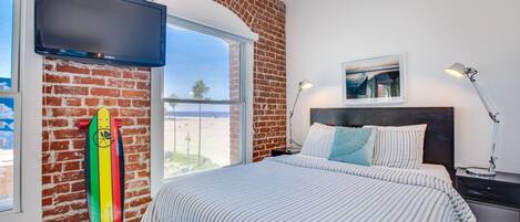 A bedroom with a brick wall, a bed with striped bedding, a mounted TV, a surfboard leaning against the wall, and a window overlooking a beach.