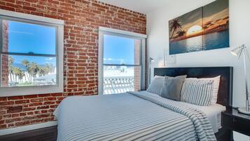 A bedroom with a brick wall, two windows, a bed with striped bedding, a nightstand, and a sunset painting on the wall.