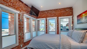 Un dormitorio con pared de ladrillo, ventanas amplias, una cama y un televisor montado en la pared.