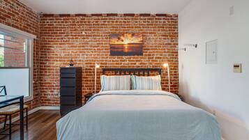Een slaapkamer met een bakstenen muur, een bed met gestreepte lakens, een bureau met stoel en een raam.