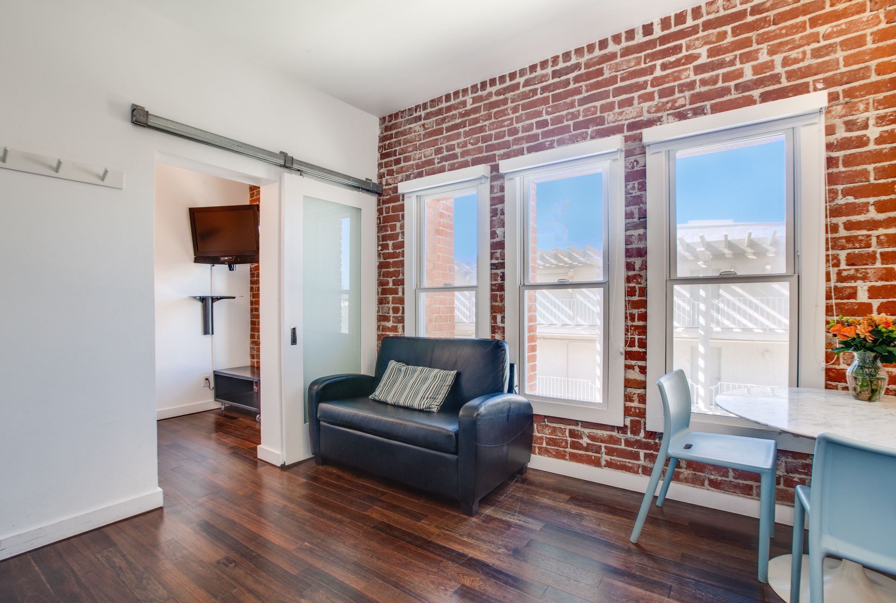 A room with a brick wall, a black leather sofa, a dining table with a chair, and a TV mounted on the wall.