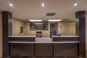 A reception desk with a man standing behind it, a bookshelf with magazines, and a door to the left. - Candlewood Suites Midwest City by IHG (Del City)