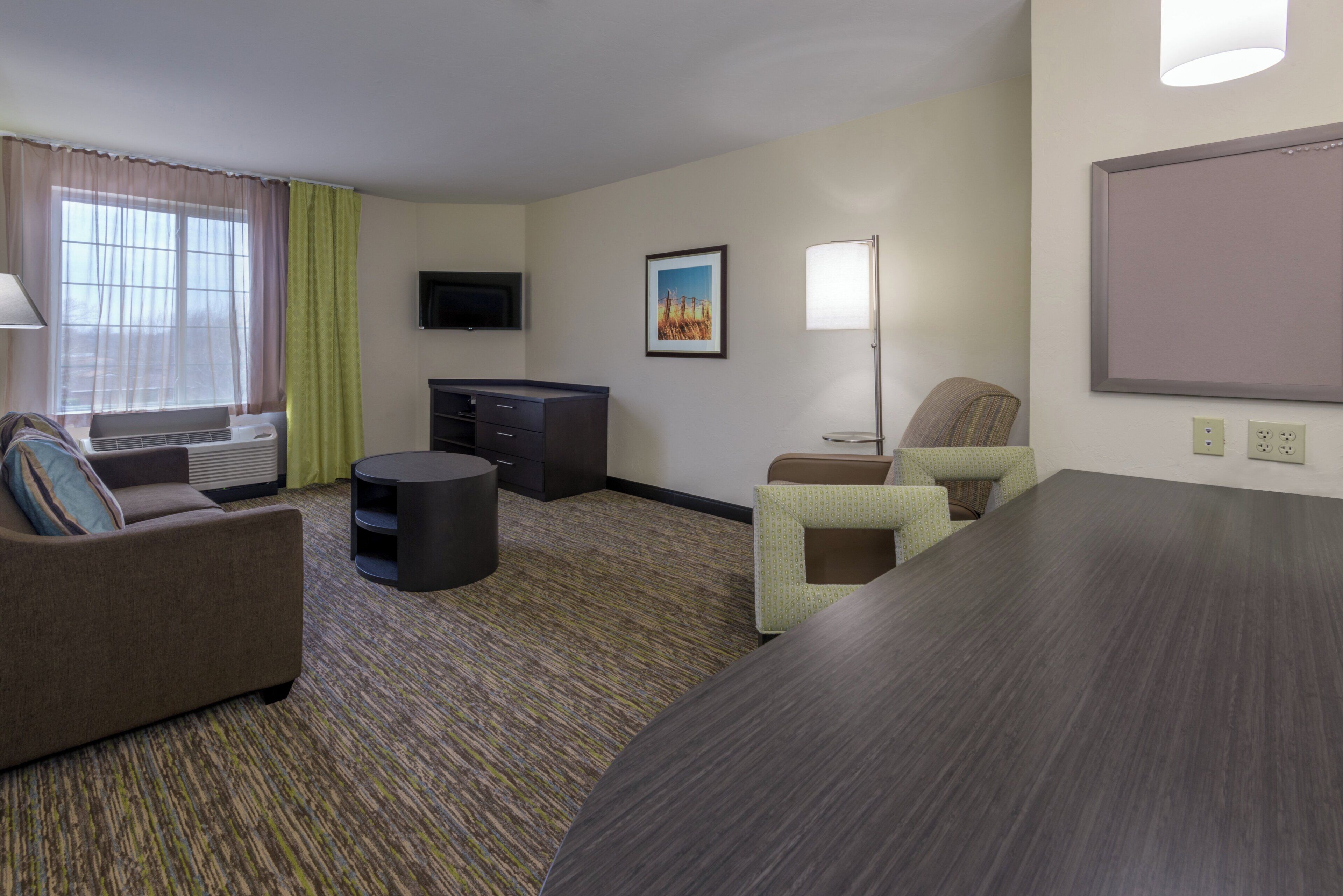 A hotel room with a sofa, a small table, a desk, a chair, a flat-screen TV, a painting, a lamp, and an air conditioning unit.