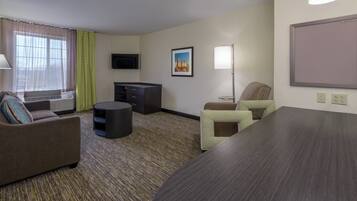 A hotel room with a sofa, a small table, a desk, a chair, a flat-screen TV, a painting, a lamp, and an air conditioning unit.