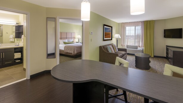 A hotel room with a bed, a sitting area with a sofa and armchairs, a dining table, a flat-screen TV, and a bathroom with a mirror and sink.