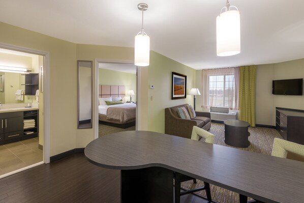 Habitación de hotel con cama, zona de estar con sofá y sillones, mesa de comedor, televisor de pantalla plana y baño con espejo y lavamanos.