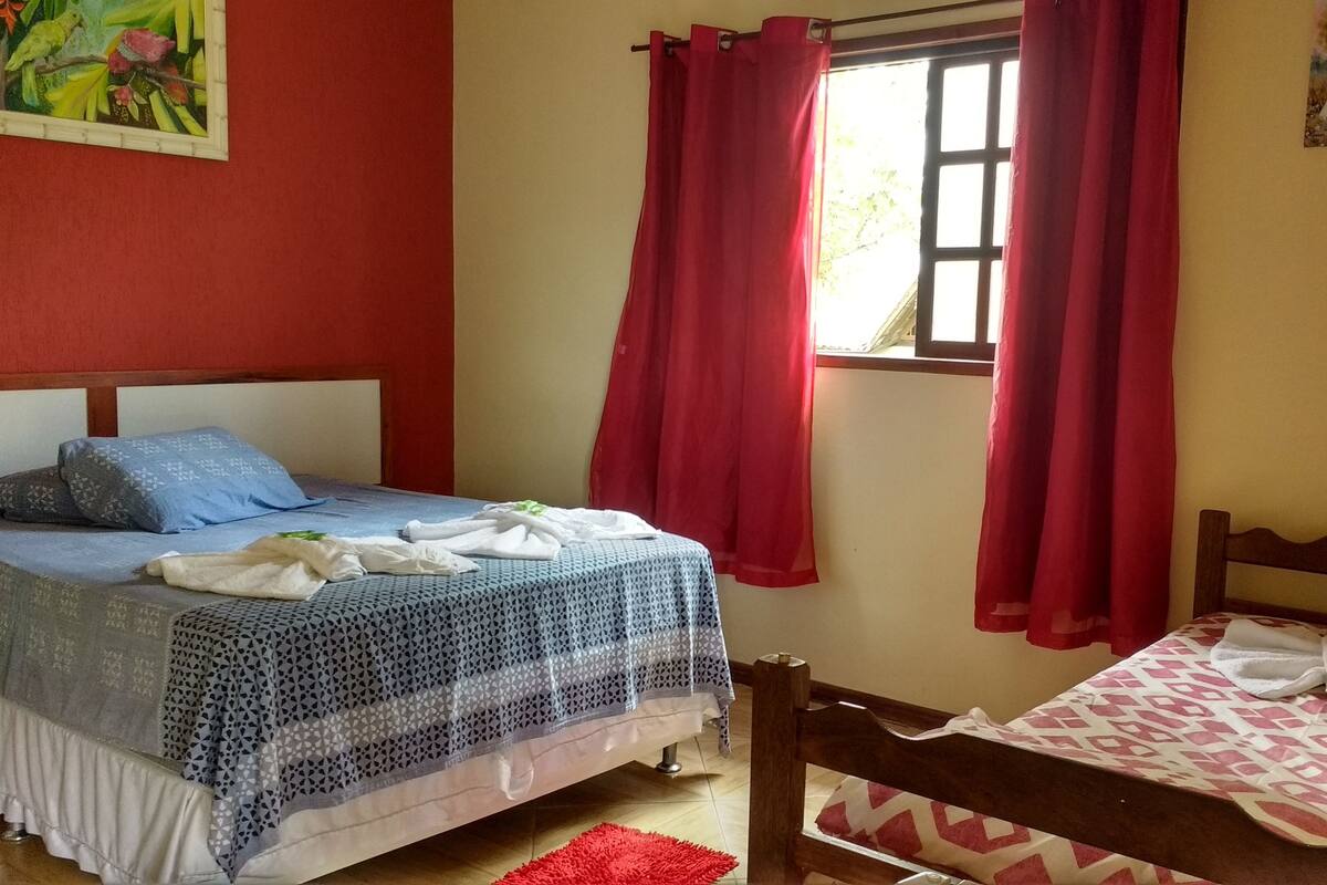 Un dormitorio con una cama, una cama individual, una ventana con cortinas rojas y un cuadro en la pared.