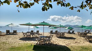 A beach with rows of wooden lounge chairs and green umbrellas, a sandy beach, and a calm sea with boats in the distance.