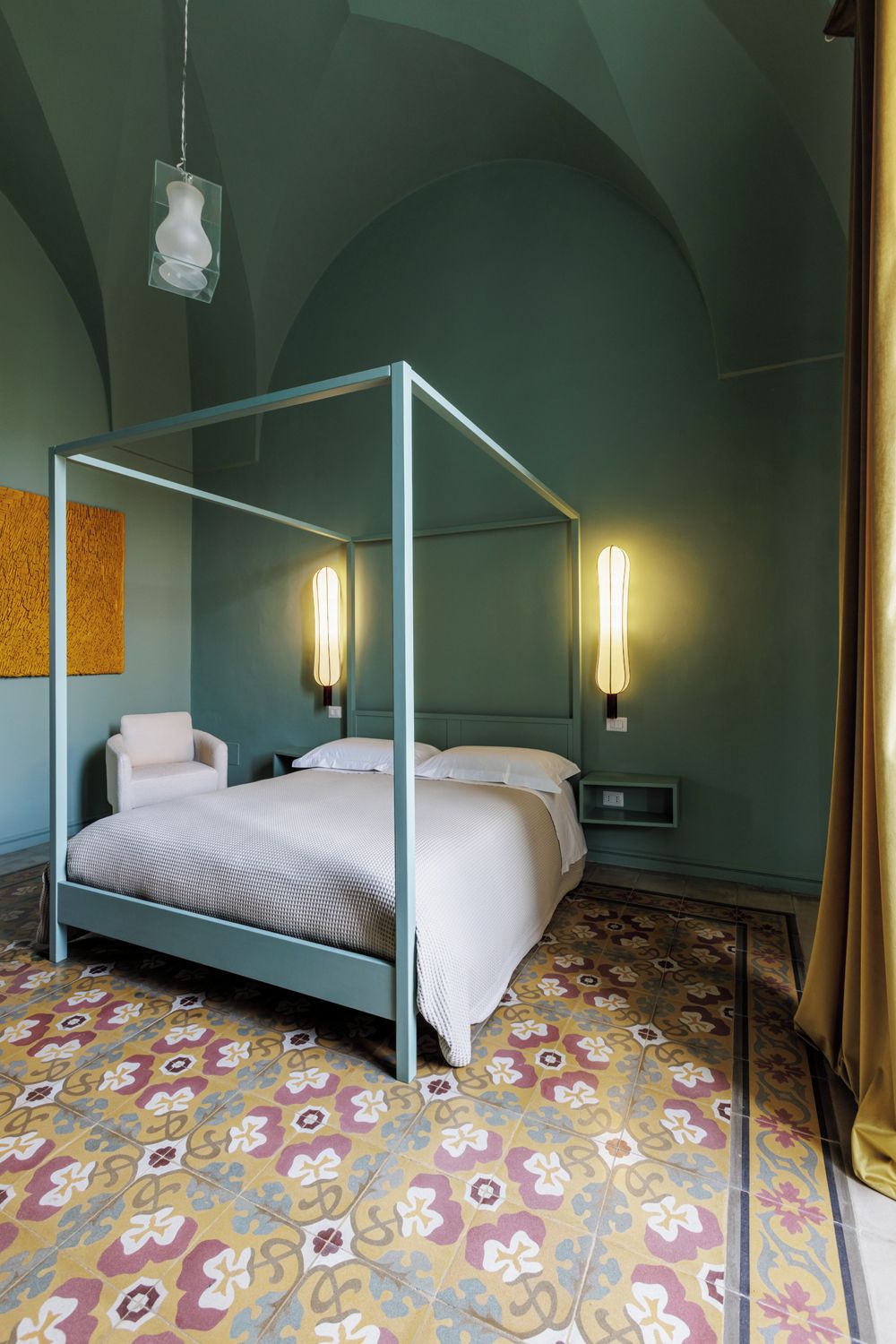 Un lit à baldaquin avec une couvre-lit blanche, une chaise et une table de chevet avec une lampe. La pièce a un mur vert et un sol à motifs.