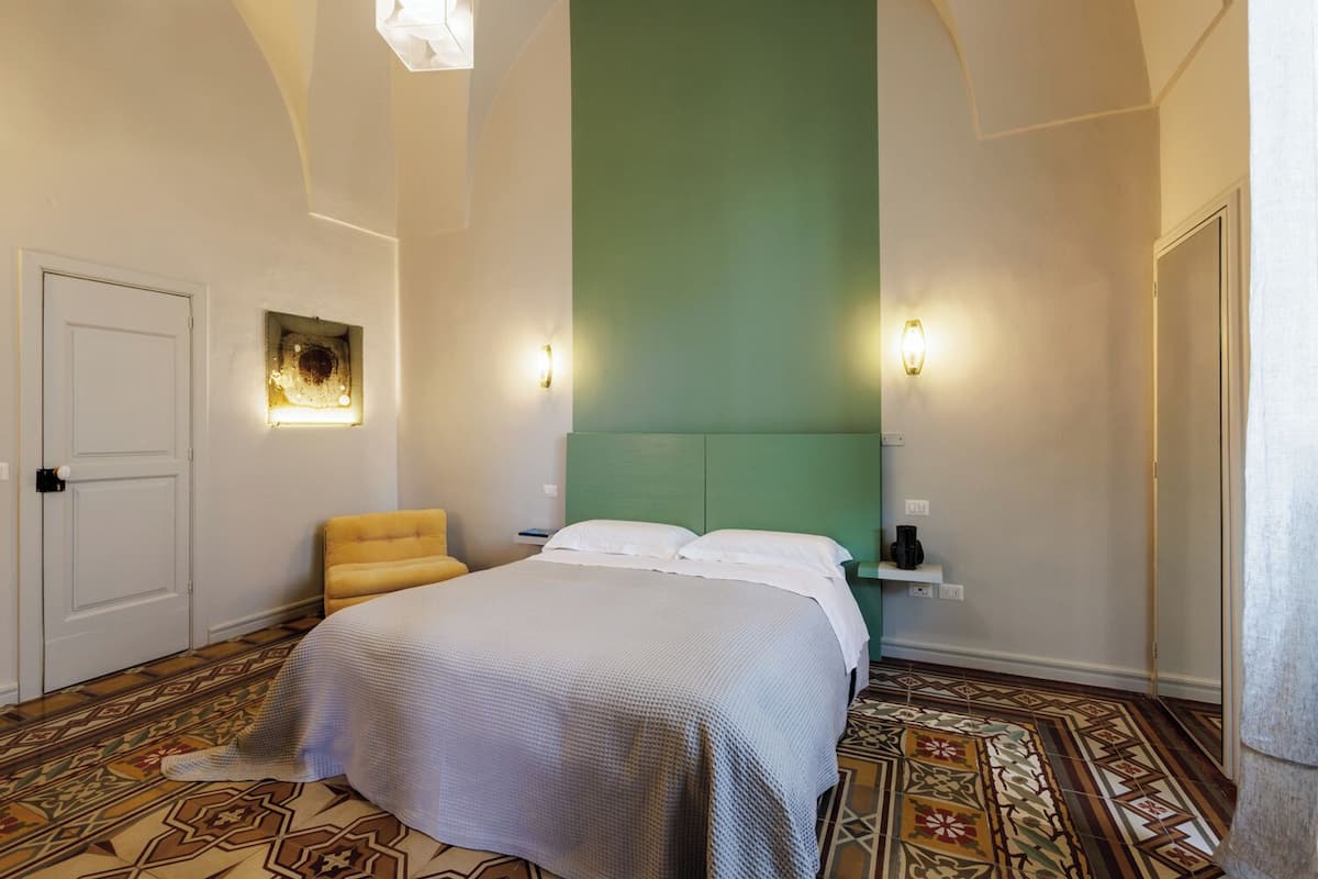 A hotel room with a bed, a chair, a green accent wall, and patterned flooring.