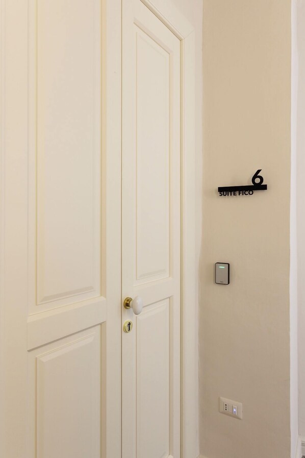 Une porte blanche avec une serrure et un trou de serrure. Une plaque noire portant le numéro 6 et l’inscription « SUITE FICO » est fixée au mur au-dessus de la porte.
