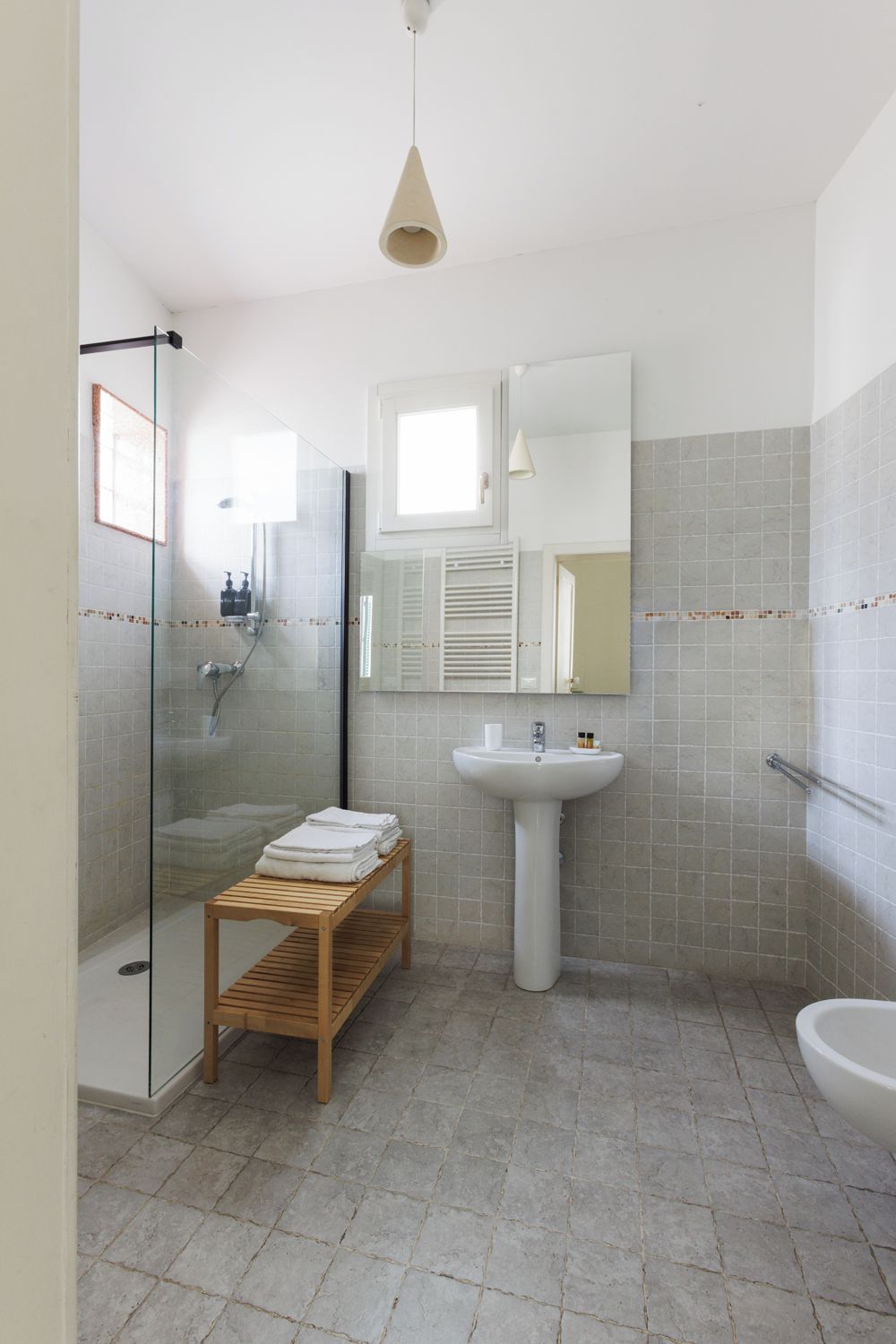Un baño moderno con ducha envuelta en vidrio, lavamanos pedestal y banco de madera.
