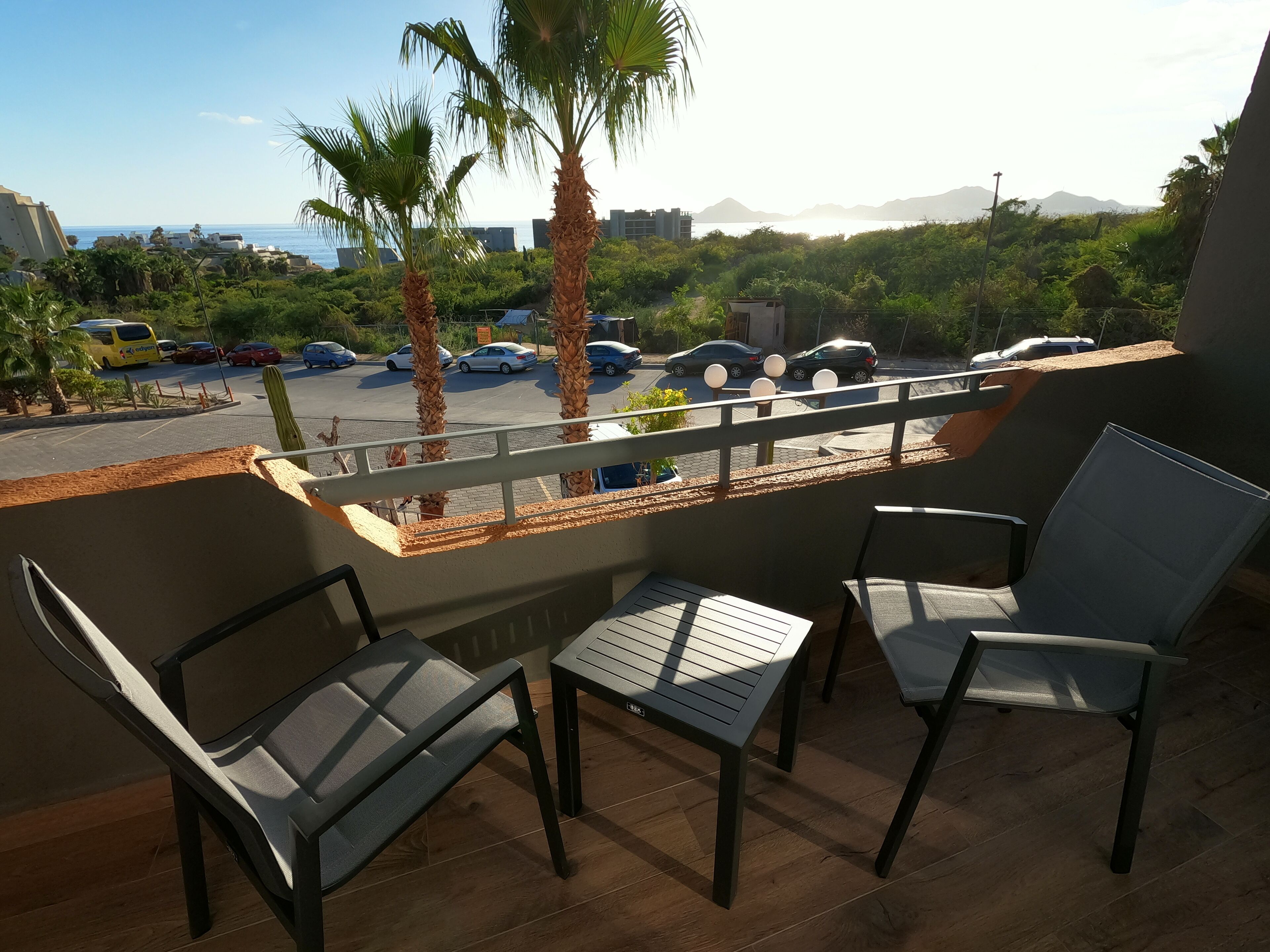Een balkon met twee stoelen en een kleine tafel, met uitzicht op een parkeerplaats met meerdere auto's en palmbomen.