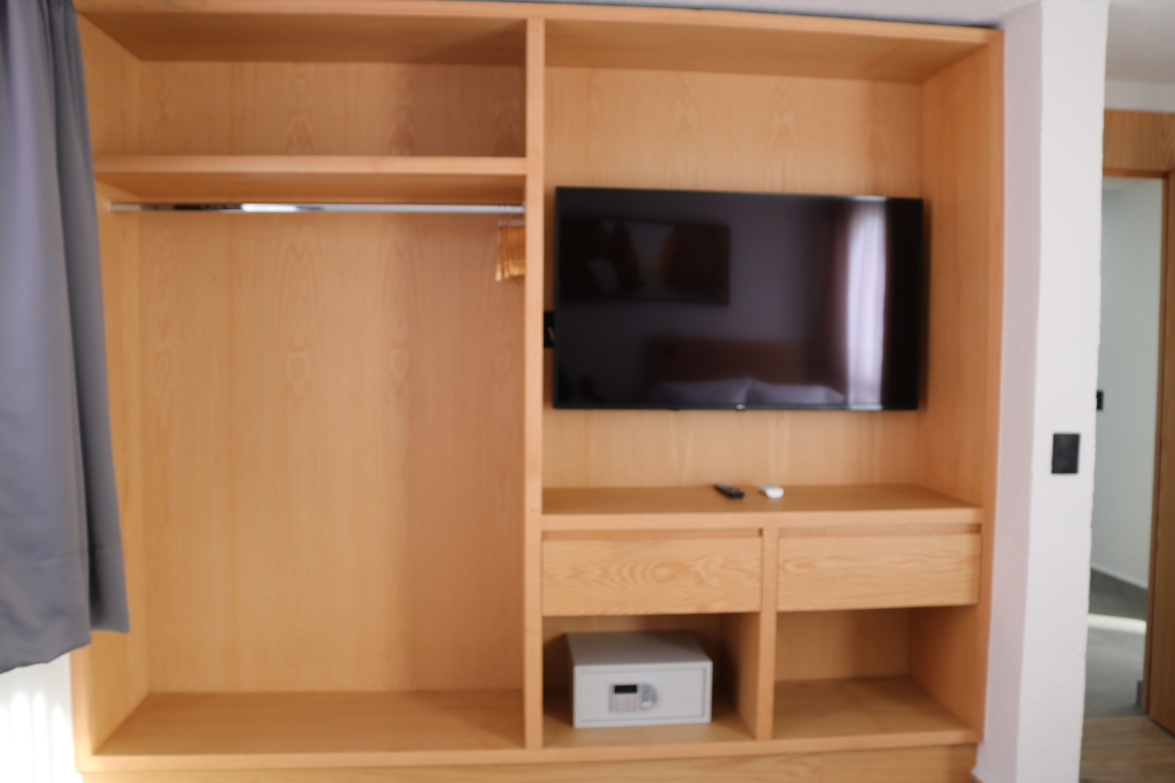 A wooden wardrobe with a flat-screen TV mounted on the door, a small drawer, and a shelf with a box.