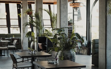 A modern restaurant interior with high ceilings, exposed concrete beams, and large windows. Tables and chairs are arranged for dining.