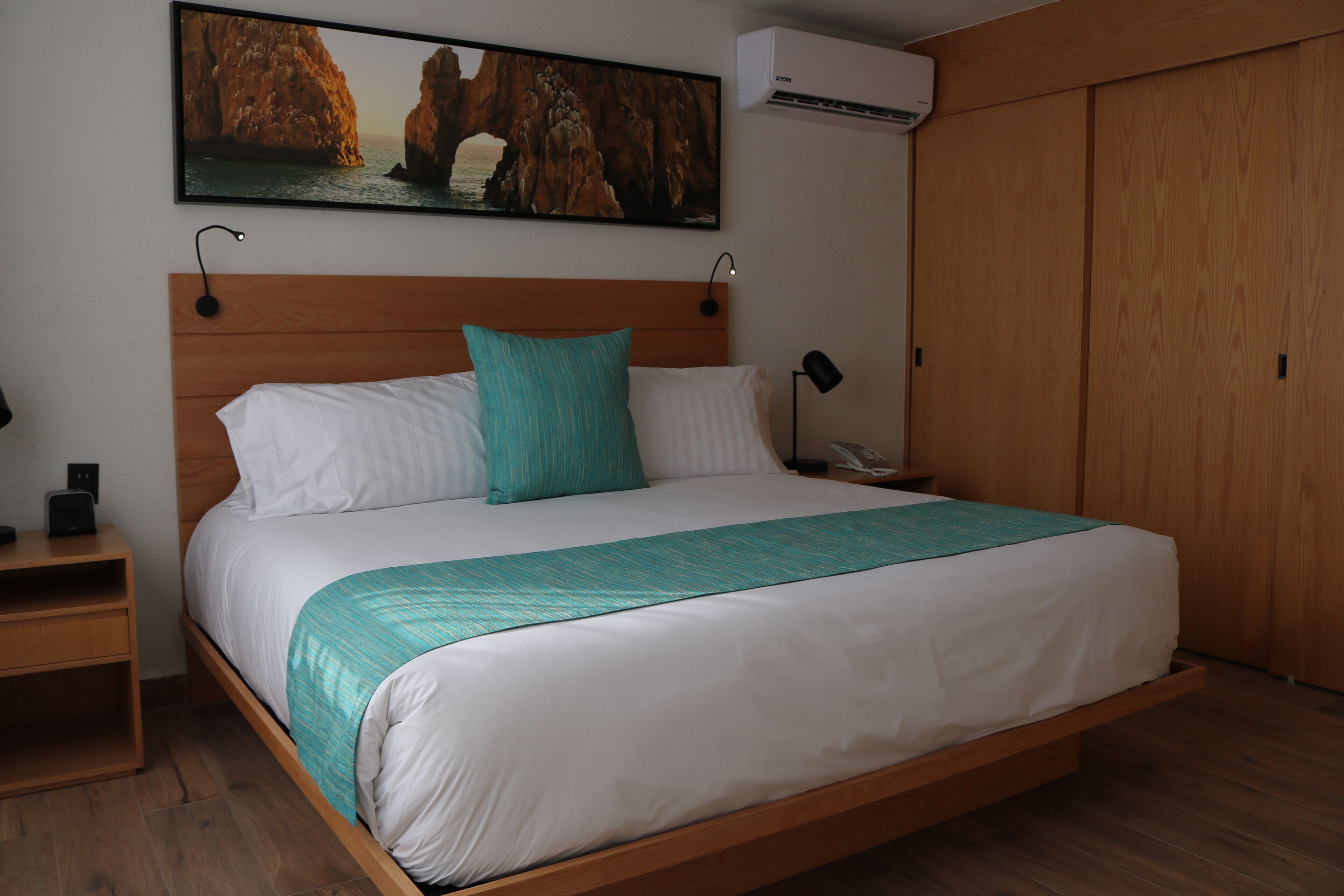 A neatly made bed with a teal blanket, wooden headboard, and a painting of a coastal scene above it.