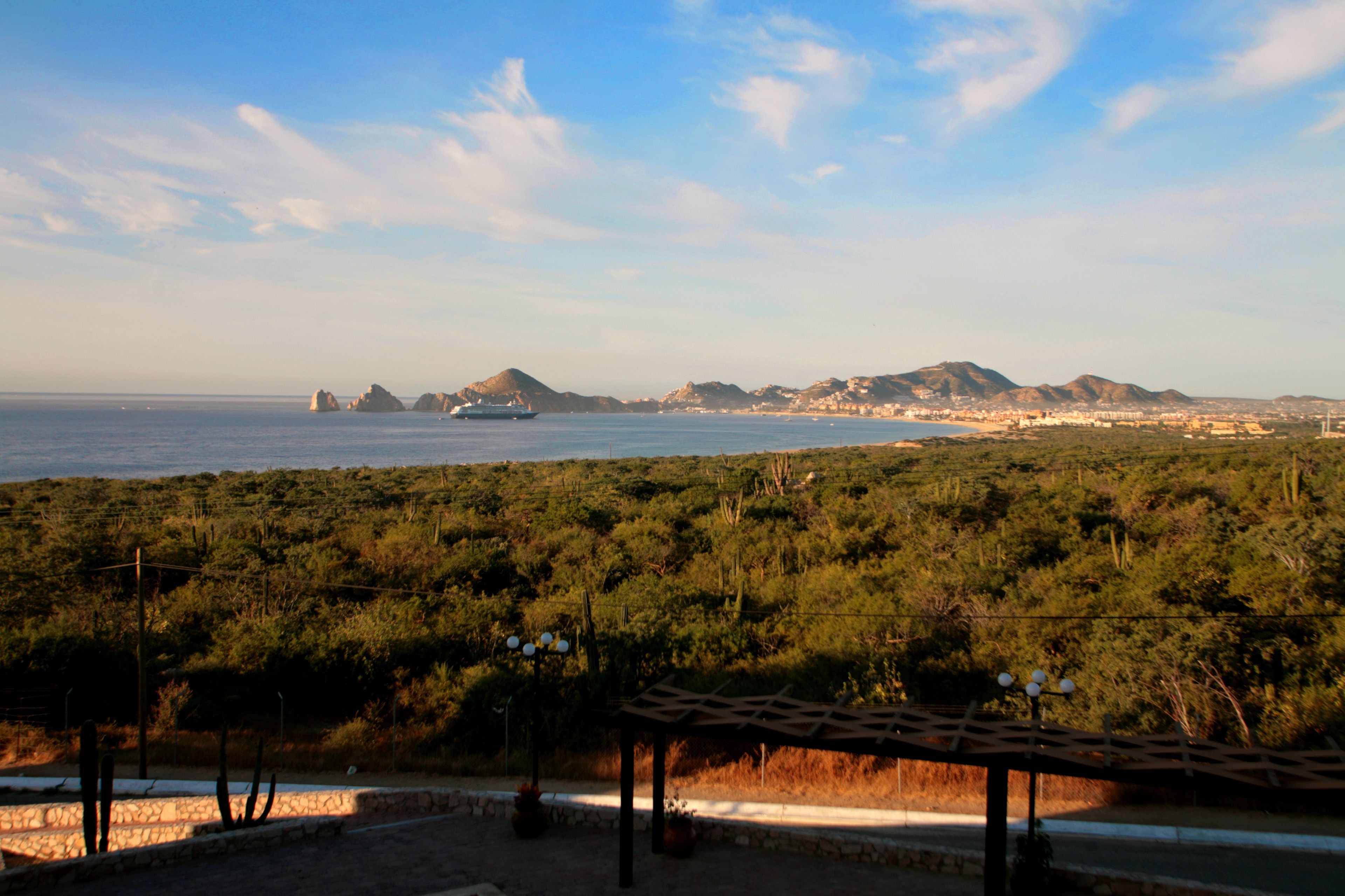 Un paysage côtier offrant une vue sur la mer, les montagnes, ainsi qu’une zone aménagée avec une pergola et des plantes en pot.