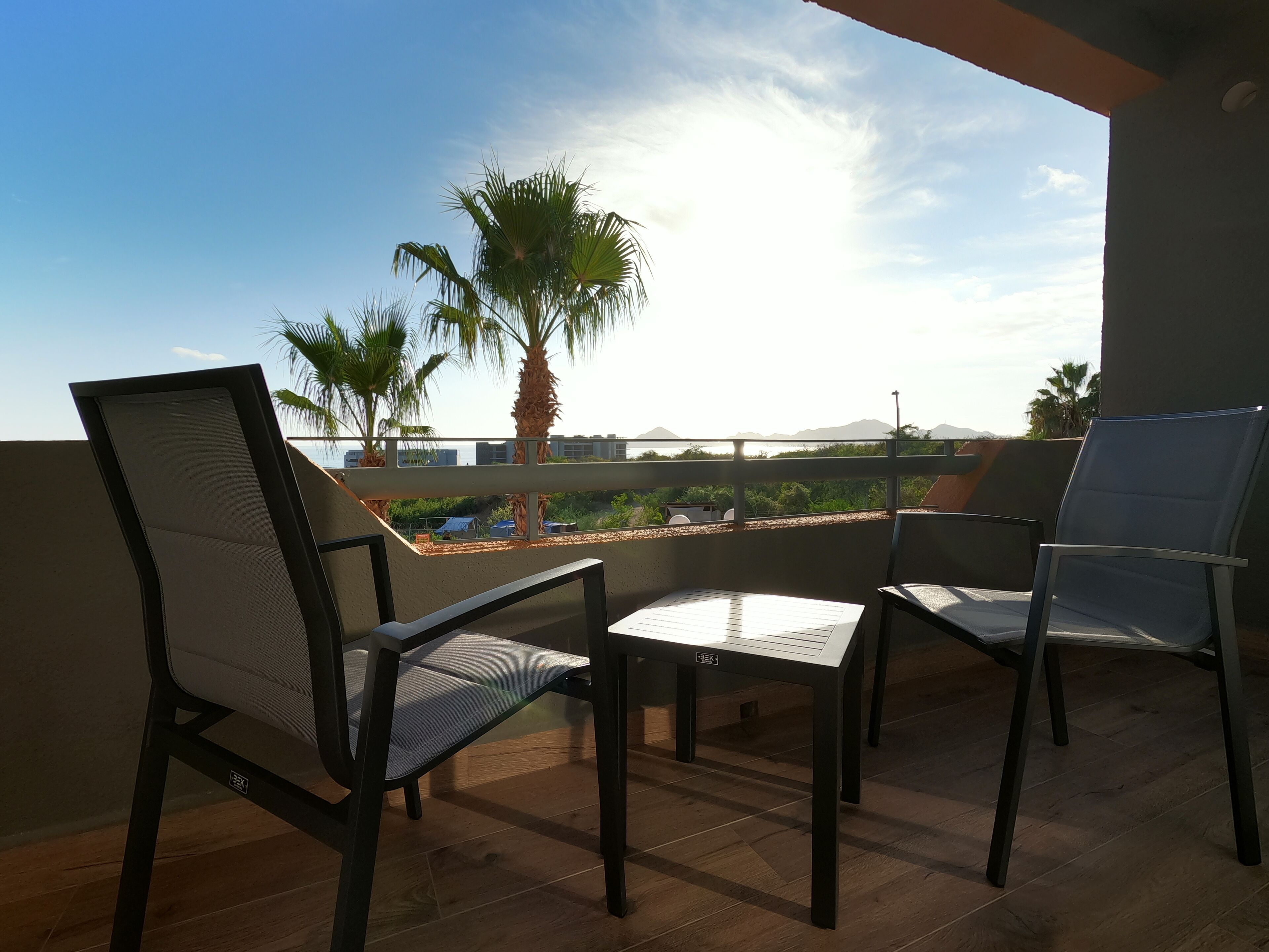 A balcony with two chairs and a table, overlooking a cityscape with palm trees and buildings.