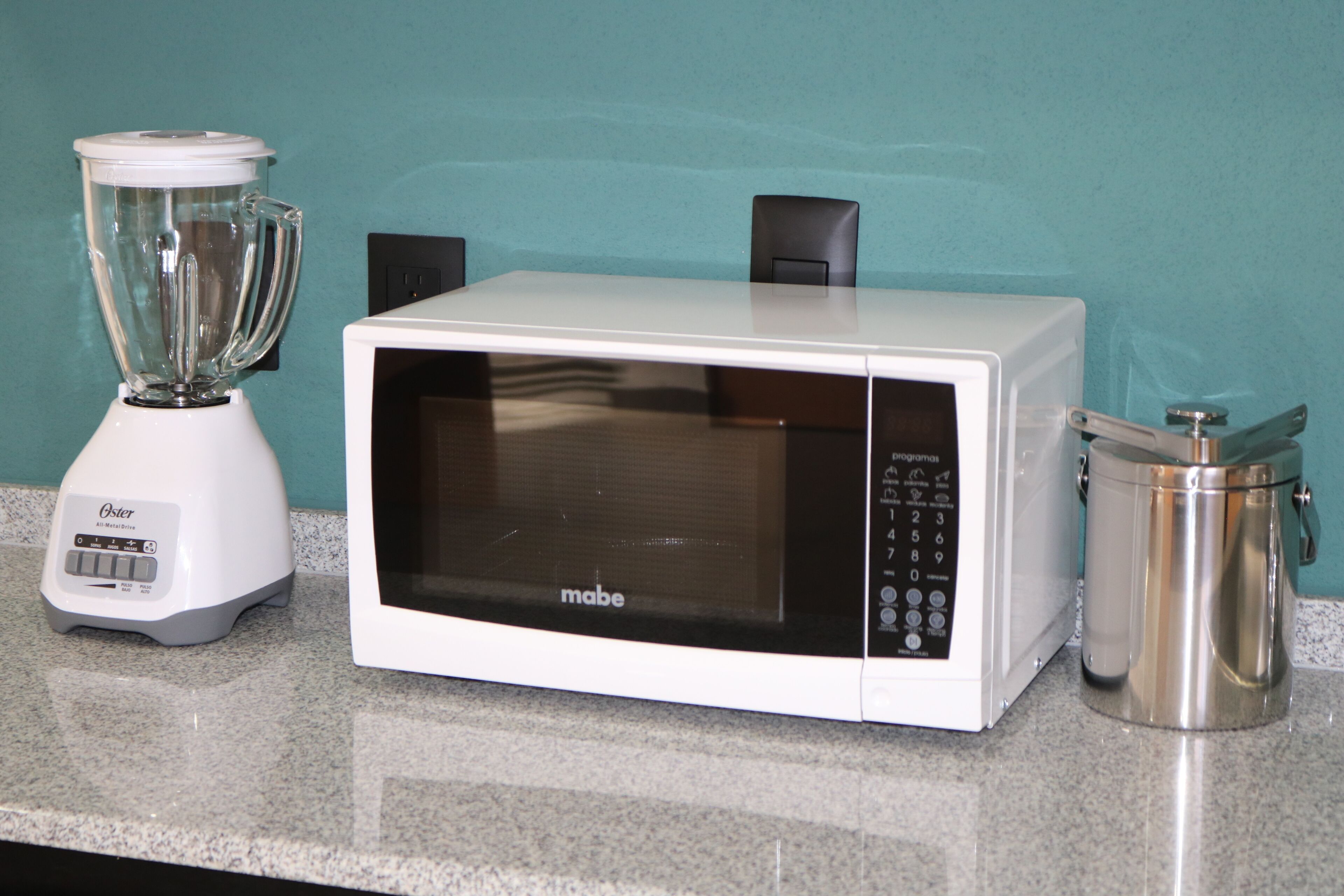 A kitchen with a white microwave, a blender, and a stainless steel container on a granite countertop.