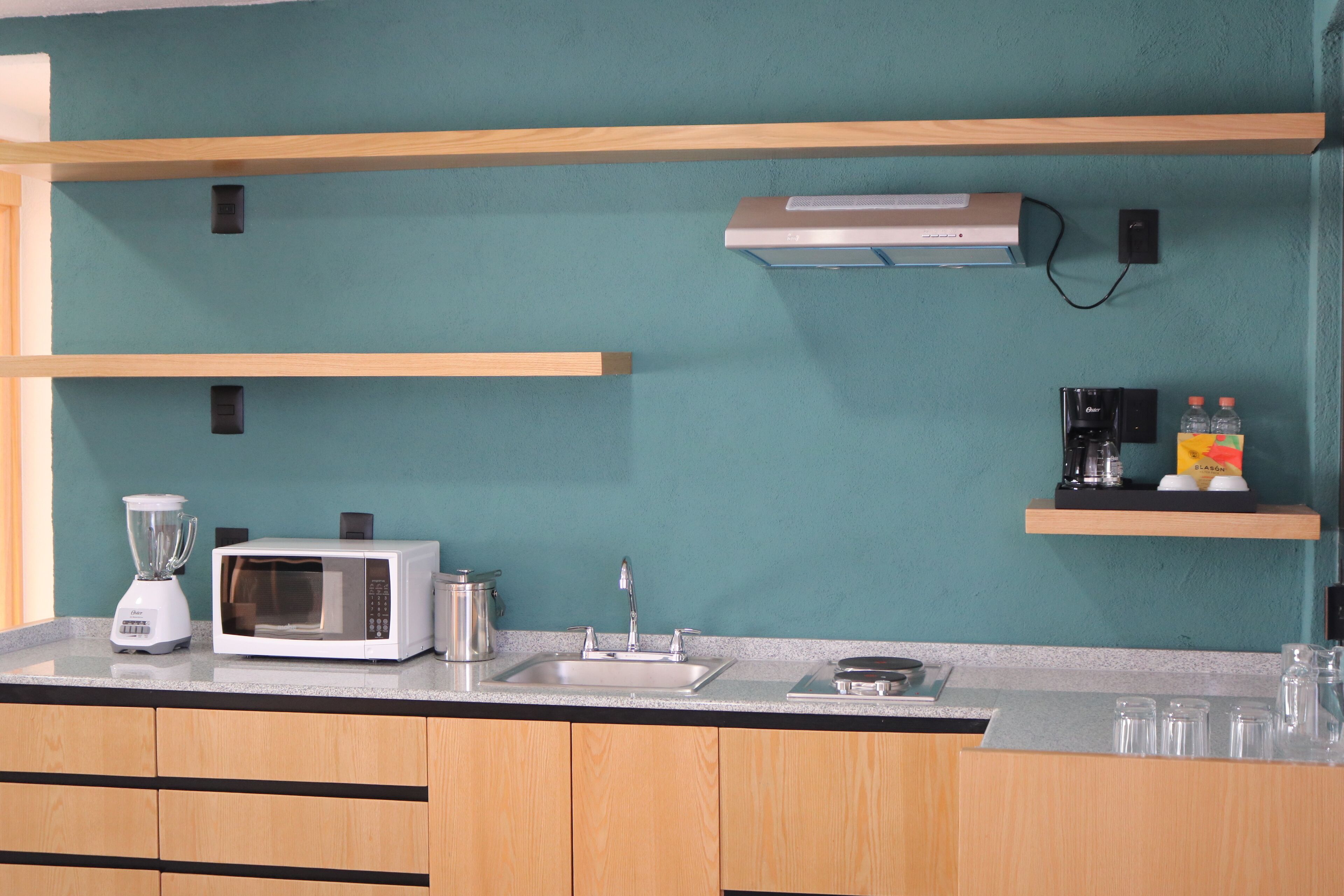 Una cocina con microondas, licuadora y lavabo. Hay repisas de madera sobre la encimera y un aire acondicionado instalado en la pared.